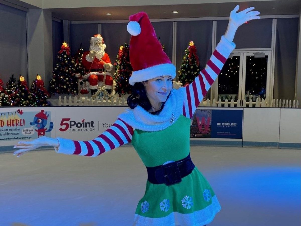 Holiday performer dressed as an elf poses on the ice rink, with Santa Claus, decorated Christmas trees, and holiday lights in the background.